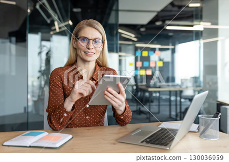 Smiling professional woman sits at a desk in an organized office, utilizing her tablet for work responsibilities while surrounded by notebooks, a laptop, and glass office walls with sticky notes. 130036959