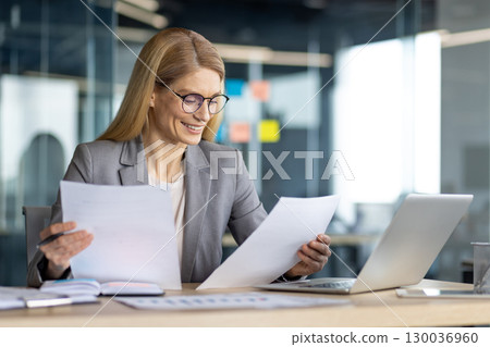 Confident businesswoman reviewing papers while working on a laptop in a bright office. 130036960