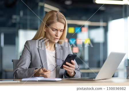 A professional woman in business attire is focused on her smartphone in a modern office setting. She appears puzzled or concerned while reviewing information, with a laptop A professional woman in business attire is focused on her smartphone in a modern office setting. She appears puzzled or concerned while reviewing information, with a laptop 130036982