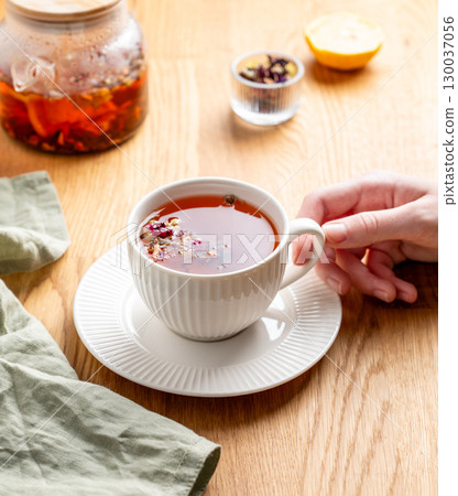 Hand holding cup of herbal tea with lemon on a wooden table with teapot and napkin. 130037056