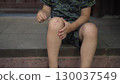 the child touches his scraped knee with a finger while sitting on the porch on a hot summer day he is wearing a t shirt and shorts 130037549