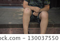 the child touches his scraped knee with a finger while sitting on the porch on a hot summer day he is wearing a t shirt and shorts 130037550