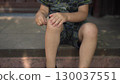 the child touches his scraped knee with a finger while sitting on the porch on a hot summer day he is wearing a t shirt and shorts 130037551