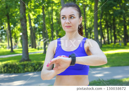 Woman checking fitness tracker while jogging in a sunny park surrounded by greenery Woman checking fitness tracker while jogging in a sunny park surrounded by greenery 130038672