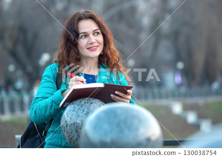 Woman writing in notebook outdoors during early evening in a park with trees 130038754