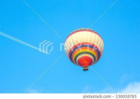Colorful hot air balloon soaring high above clear blue sky with jet plane trail 130038765