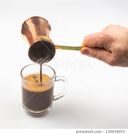 A hand pours rich Turkish coffee from a copper cezve into a transparent glass cup A hand pours rich Turkish coffee from a copper cezve into a transparent glass cup 130039053