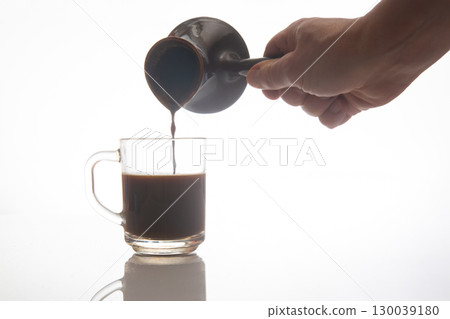 A hand pours rich coffee from a traditional ceramic pot into a clear glass cup 130039180