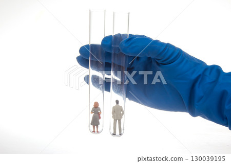 A gloved hand holds two test tubes containing miniature figurines of a man and woman 130039195