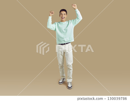 Young funny boy do winner gesture clenching fists celebrating success on beige background Young funny boy do winner gesture clenching fists celebrating success on beige background 130039786