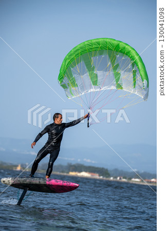 A middle-aged man in a wetsuit glides on a foil board across the water, holding a large green and white wing under a clear sky 130041098
