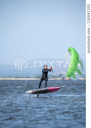 A middle-aged man in a wetsuit stands on a hydrofoil board with a green and white kite A middle-aged man in a wetsuit stands on a hydrofoil board with a green and white kite 130041103