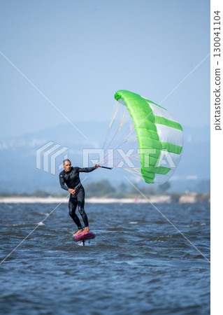 A middle-aged man in a wetsuit glides on a hydrofoil board with a green and white kite 130041104