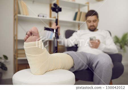 Man with bandaged leg using mobile phone while sitting in armchair 130042012