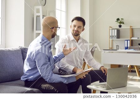 Two young businessmen having a discussion while sitting on the sofa in the office Two young businessmen having a discussion while sitting on the sofa in the office 130042036