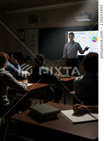 A teenage boy, 16, presents a project on a projector screen to a dimly lit classroom of students in their late teens, all focused on his presentation 130043443
