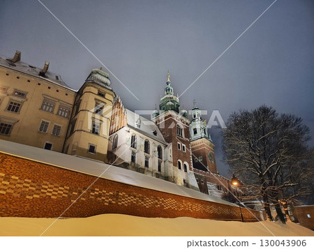 Snow covers the ground in front of an illuminated, historic building with multiple spired towers, a part of a Krakow castle 130043906