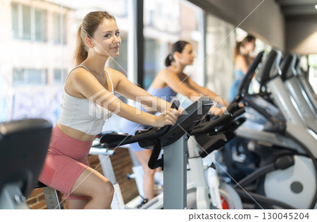 Smiling young girl doing spin bike exercises in well-equipped gym Smiling young girl doing spin bike exercises in well-equipped gym 130045204