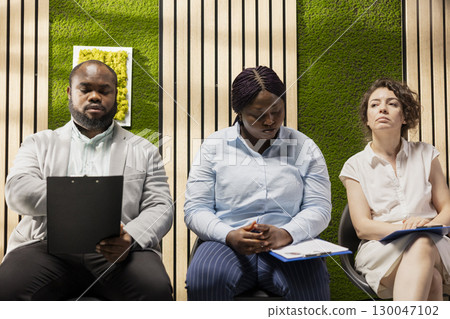 Professional diverse candidates awaiting start of job interview, showcasing anticipation in office hallway, preparing with information and CVs for the recruitment process. Career opportunity. 130047102