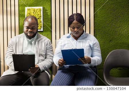 African american job seekers waiting in the corridor with their resumes, preparing for a formal interview with the HR manager. People reflecting nervousness and confidence during head hunting. 130047104