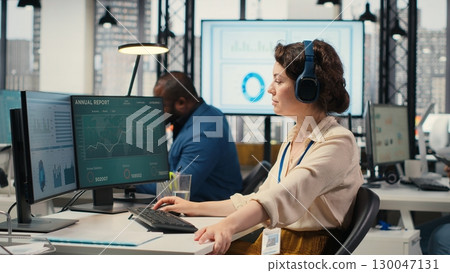 Office administrator uses focus music on headset to solve tasks, monitoring performance metrics at the workstation. Administrative tasks to ensure enterprise productivity and success. Camera B. Office administrator uses focus music on headset to solve tasks, monitoring performance metrics at the workstation. Administrative tasks to ensure enterprise productivity and success. Camera B. 130047131