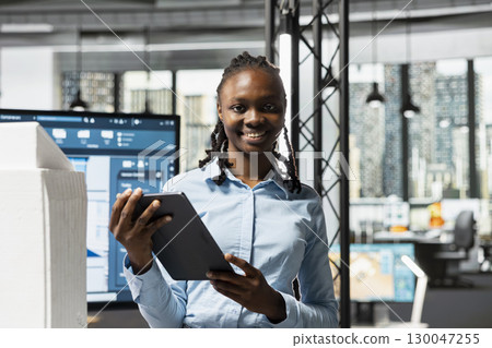 Portrait of african american woman studies blueprints on tablet for construction, smart city apartment site preparation. Engineer preparing for housing project building with 3D models. 130047255