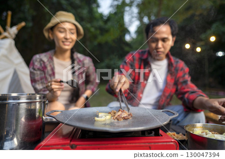 Indonesian southeast asian couple with tongs cooking sliced marinated beef bulgogi on a round grill pan with onions. The scene of the lifestyle of enjoying food in a natural setting 130047394