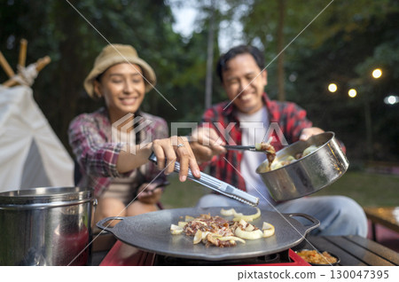Indonesian southeast asian couple with tongs cooking sliced marinated beef bulgogi on a round grill pan with onions. The scene of the lifestyle of enjoying food in a natural setting 130047395