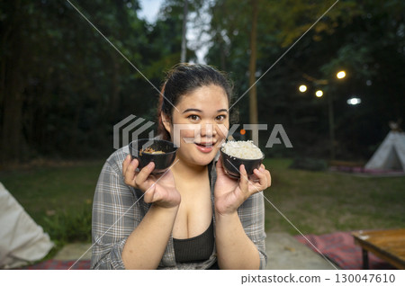 Indonesian southeast asian overweight woman holding a bowl of rice and a barbecue meal while glamping. The scene of the lifestyle of enjoying food in a natural setting Indonesian southeast asian overweight woman holding a bowl of rice and a barbecue meal while glamping. The scene of the lifestyle of enjoying food in a natural setting 130047610