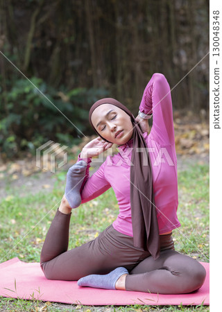 Indonesian southeast asian muslim woman in a sporty outfit practices yoga on a fitness mat in a public park outdoors. Concept of a Healthy active lifestyle 130048348