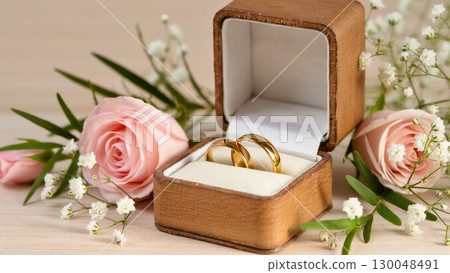 Two golden wedding rings in a leather box surrounded by pink roses and white flowers, symbolizing love, union and romantic commitment. 130048491