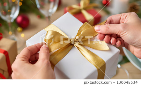 Christmas gift wrapping with golden ribbon in female hands, festive holiday scene with blurred decorations in background 130048534
