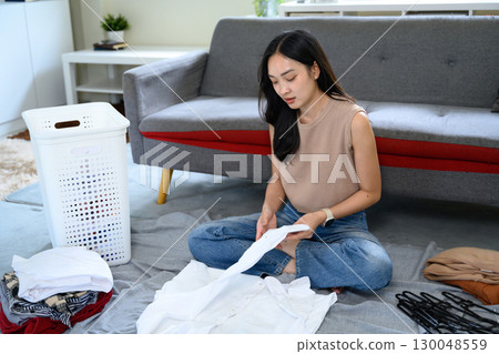Young woman folding clean laundry at home, sitting on floor with clothes and basket 130048559