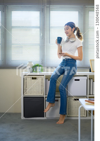 Young woman holding coffee mug and smartphone while relaxing at home near window 130048568