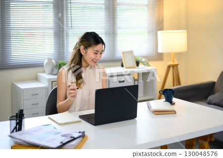 Smiling young Asian woman working from home with laptop 130048576