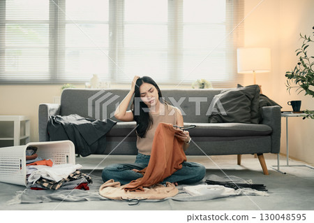 Frustrated woman sitting on floor with laundry basket, stressed while folding clothes at home 130048595