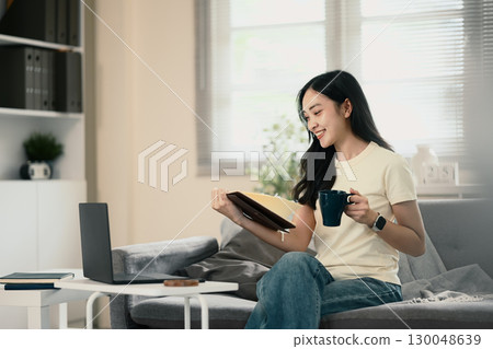 Smiling woman holding notebook and coffee mug while working from home 130048639