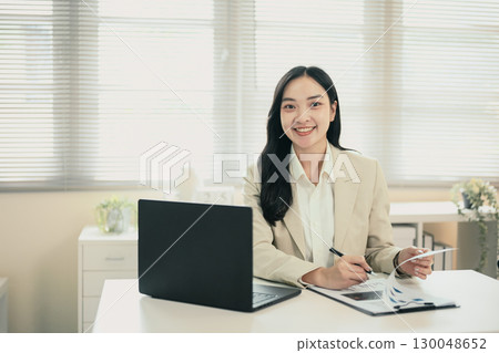 Professional businesswoman smiling at desk with laptop and documents in modern office Professional businesswoman smiling at desk with laptop and documents in modern office 130048652