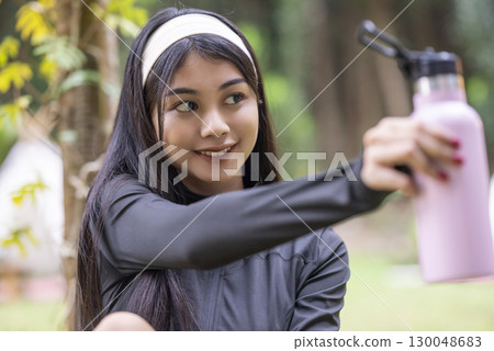 Indonesian southeast asian woman in a sporty outfit drinking water after practice running exercise in a public park outdoors. Concept of a Healthy active lifestyle 130048683