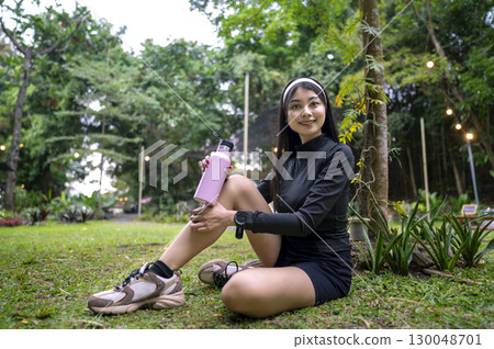 Indonesian southeast asian woman in a sporty outfit holding a drinking bottle after practice running exercise in a public park outdoors. Concept of a Healthy active lifestyle Indonesian southeast asian woman in a sporty outfit holding a drinking bottle after practice running exercise in a public park outdoors. Concept of a Healthy active lifestyle 130048701