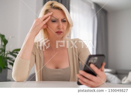 Middle aged blonde woman sitting in modern cozy interior, holding phone and looking at screen with worried and puzzled expression, concept of unexpected news or problems 130049827