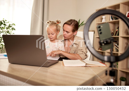 Mother assisting young daughter with homework at home using laptop and books Mother assisting young daughter with homework at home using laptop and books 130050432
