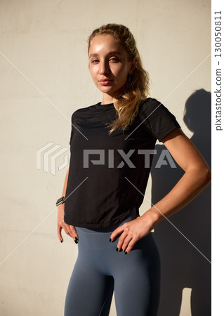 Confident woman in activewear posing against a wall in natural sunlight Confident woman in activewear posing against a wall in natural sunlight 130050811