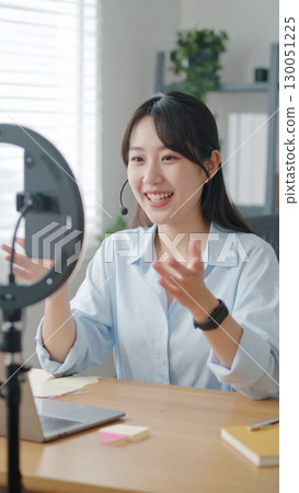 一位年輕女子正在家庭辦公室使用環形燈和智慧型手機，一邊微笑一邊直播網路研討會或召開線上會議。 130051225