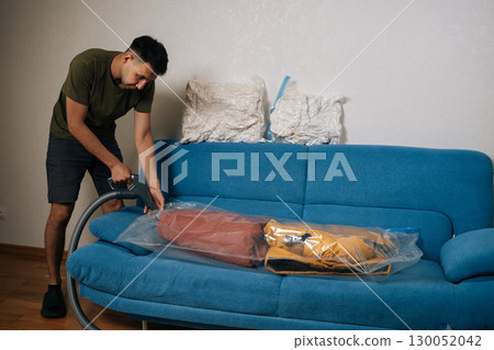 Man using vacuum cleaner to remove air from vacuum storage bag with clothes and placing it on blue sofa in living room, organizing seasonal wardrobe for winter. Concept of household chores. Man using vacuum cleaner to remove air from vacuum storage bag with clothes and placing it on blue sofa in living room, organizing seasonal wardrobe for winter. Concept of household chores. 130052042