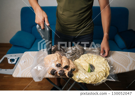 Cropped shot of man using vacuum cleaner to suck air out of plastic bags containing plush dog toys, placed on ironing board, for efficient storage and space saving. Concept of household chores. 130052047