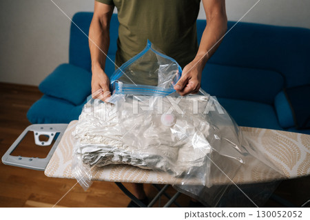 Unrecognizable housewife male closing vacuum storage bag placed on ironing board, containing bedding or out-of-season clothes, sealing for efficient space-saving storage. Concept of household chores. 130052052