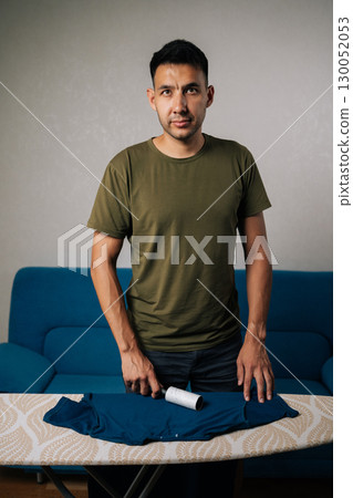 Vertical portrait of handsome man removing lint from blue t-shirt using sticky roller, preparing clothing for pressing, looking at camera. Concept of domestic work, housekeeping and household. 130052053