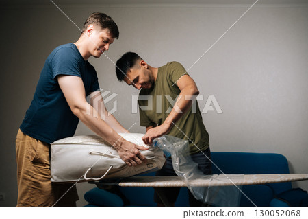 Two young men working together, using vacuum cleaner to pack bedding into vacuum storage bag placed on ironing board, efficiently organizing living space at home. Concept of household chores. 130052068