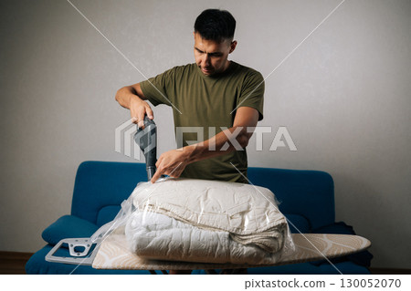 Front view of man using vacuum cleaner to extract air from storage bag filled with bedding, positioned on ironing board, creating efficient space-saving storage solution. Concept of household chores. 130052070